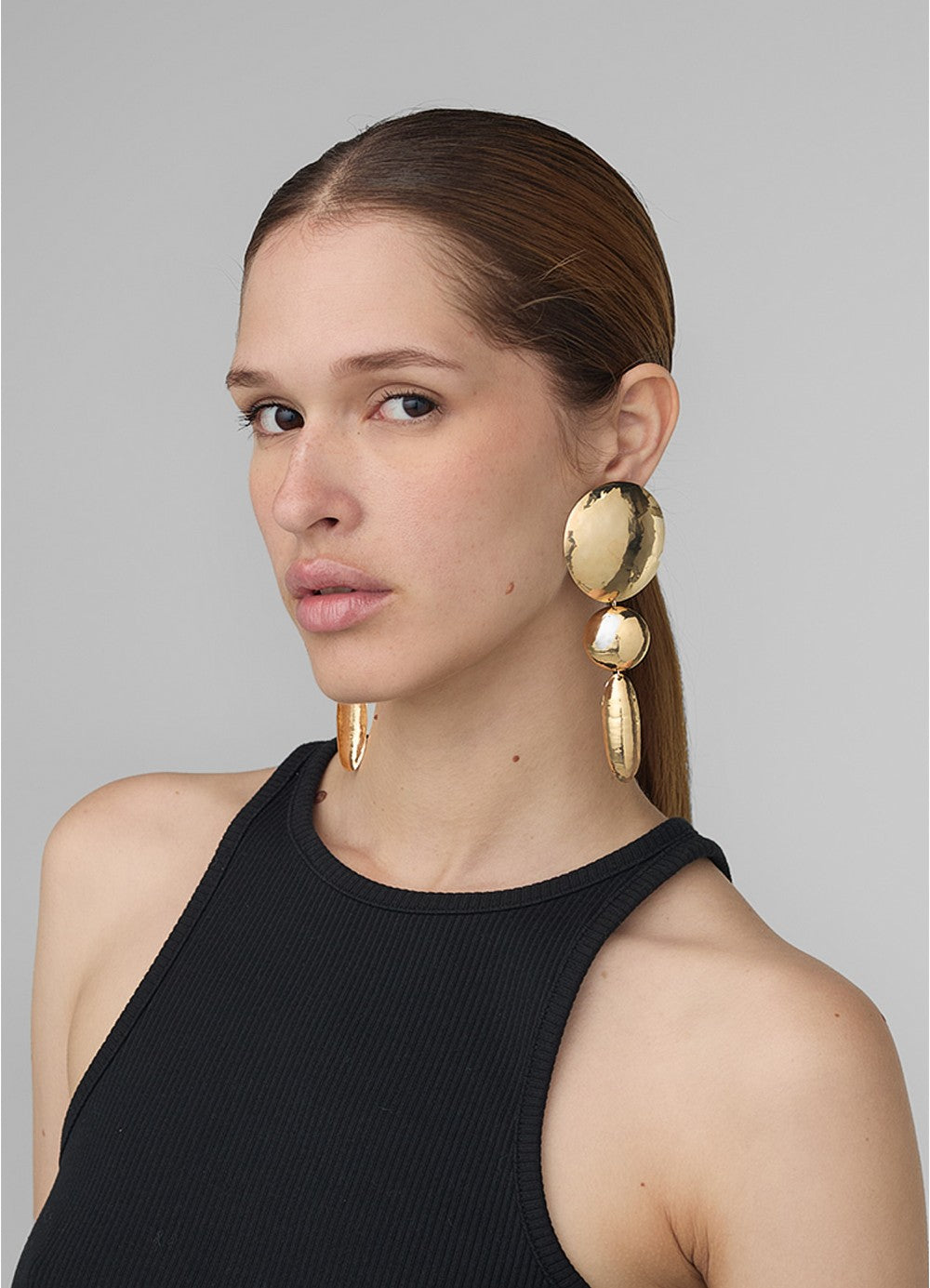 Modelo con look pulido usando aretes grandes y largos en dorado compuestos por dos círculos y un
óvalo, todos con textura repujada; una pieza de diseño escultórico de Paula Mendoza.