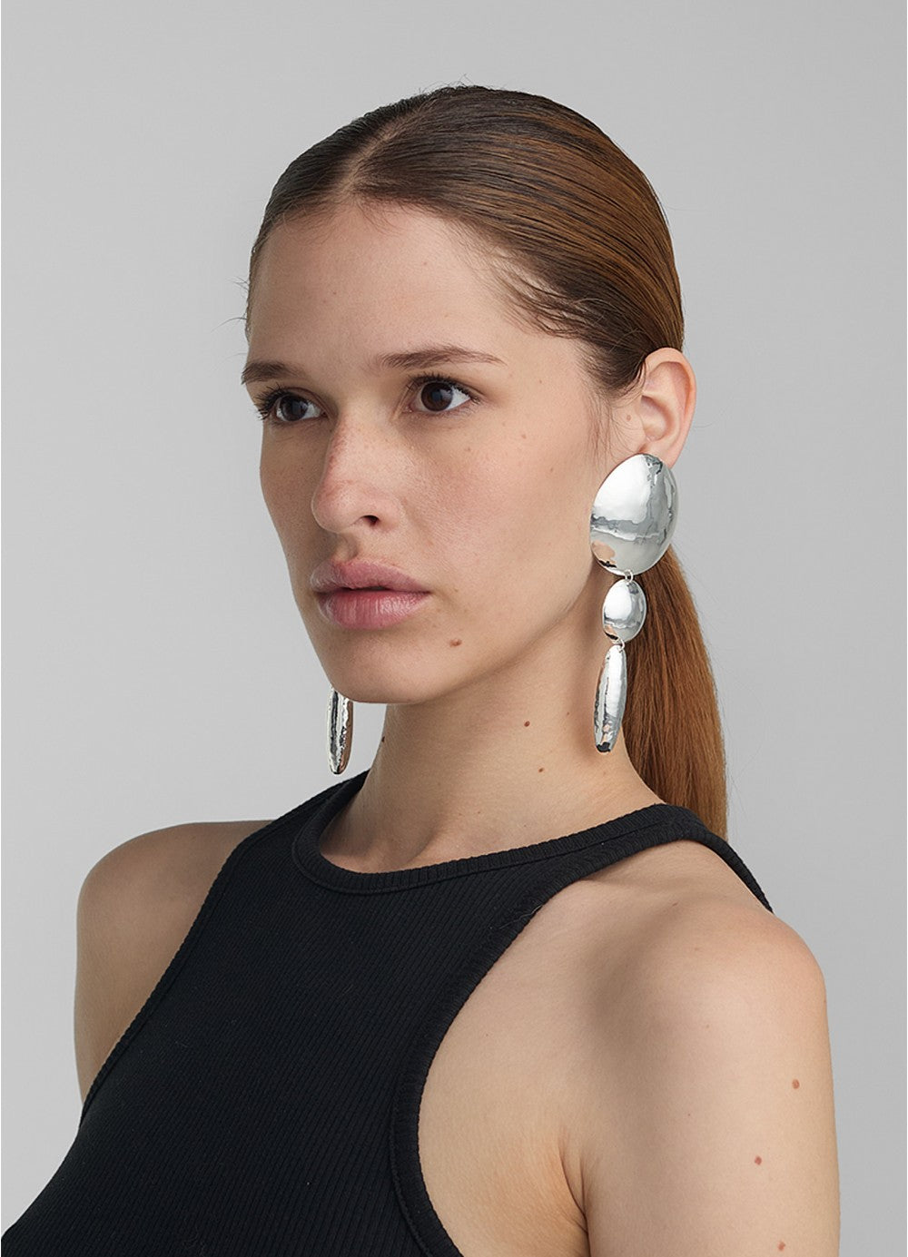 Modelo con look pulido usando aretes grandes y largos en plateado compuestos por dos círculos y un
óvalo, todos con textura repujada; una pieza de diseño escultórico de Paula Mendoza.